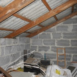VSL Szymon Lendzion - Surowe poddasze z drewnianą konstrukcją dachu i ścianami z szarych bloczków. Widoczne elementy instalacji i akcesoria budowlane. Nierówności i niedokończone wykończenie.