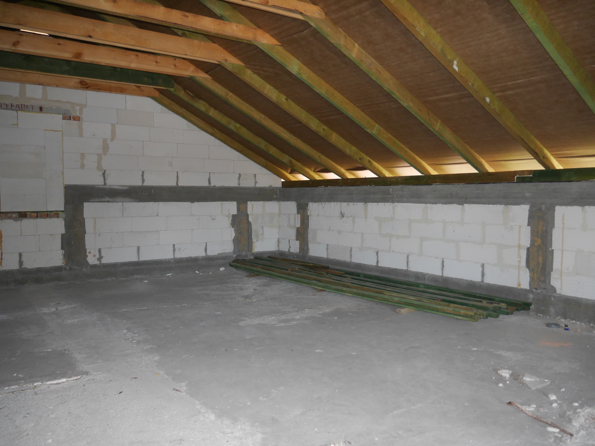 Surowe poddasze z widoczną konstrukcją dachu, ścianami z betonu komórkowego i stertą zielonych łat. Wnętrze w trakcie budowy lub remontu.