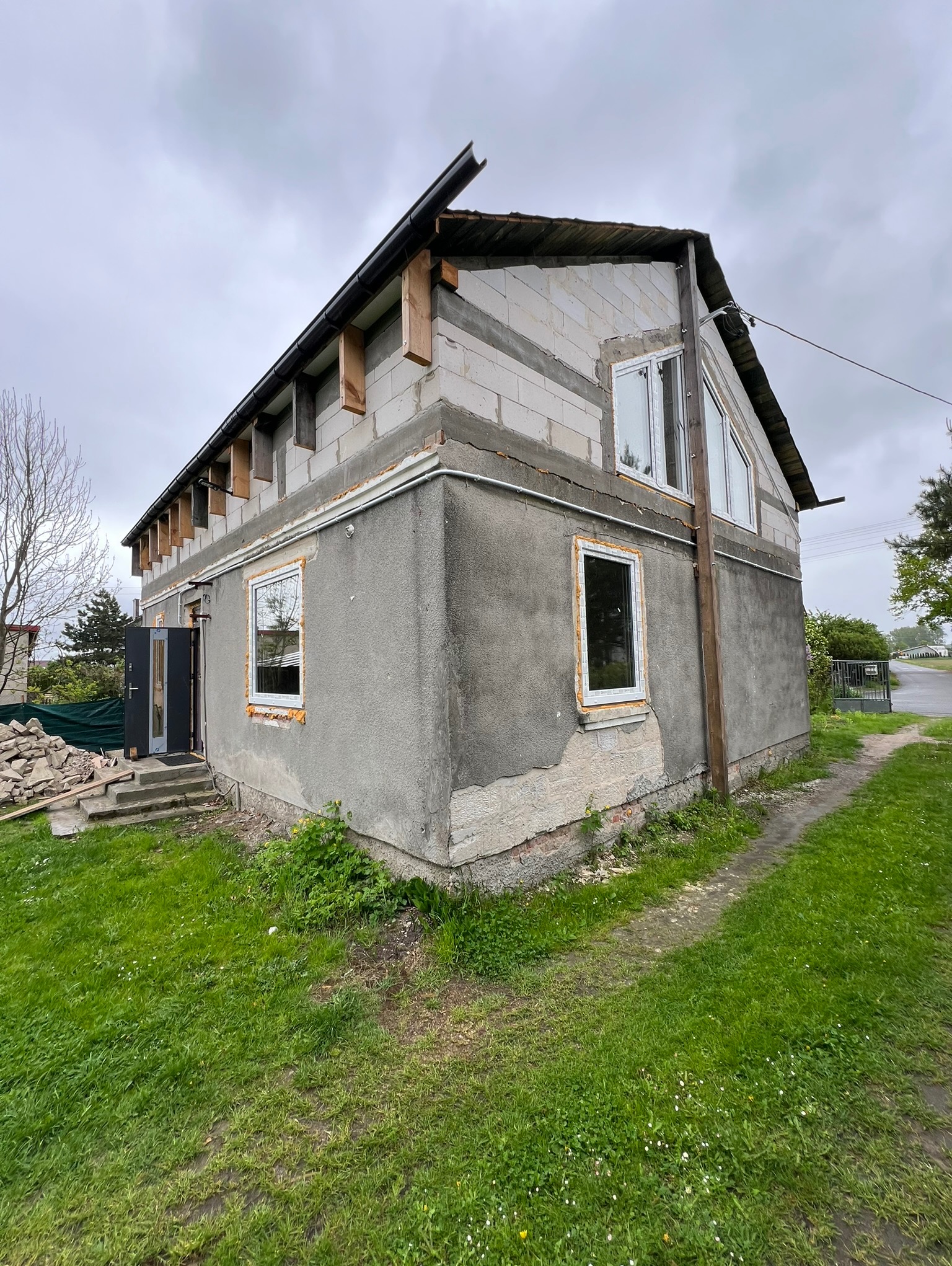 Dom w trakcie budowy/remontu: widoczne okna, surowy tynk, bloczki, elementy konstrukcji dachu. Otoczenie: trawa, ścieżka, pochmurne niebo.