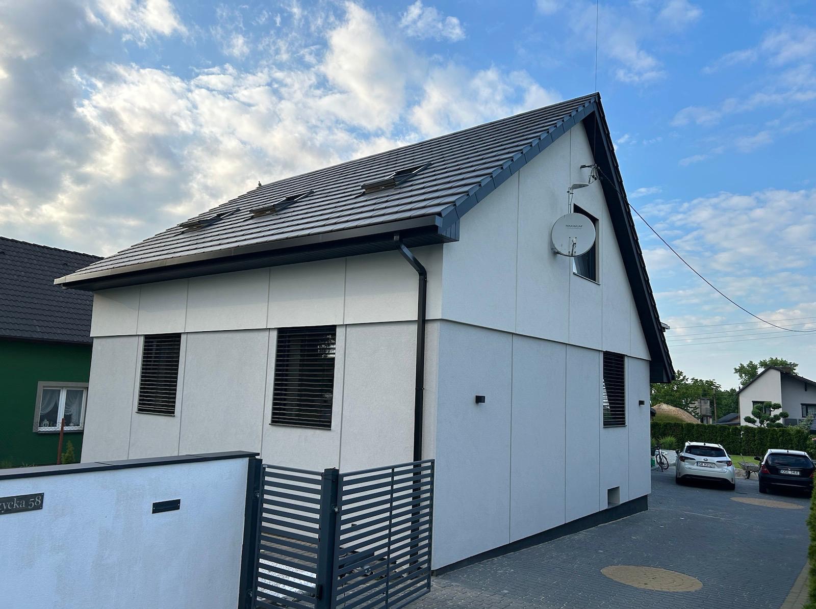 Nowoczesny dom jednorodzinny z szarą elewacją i ciemną dachówką, widok z zewnątrz na tle błękitnego nieba z chmurami. Widoczne detale: antena satelitarna, żaluzje.