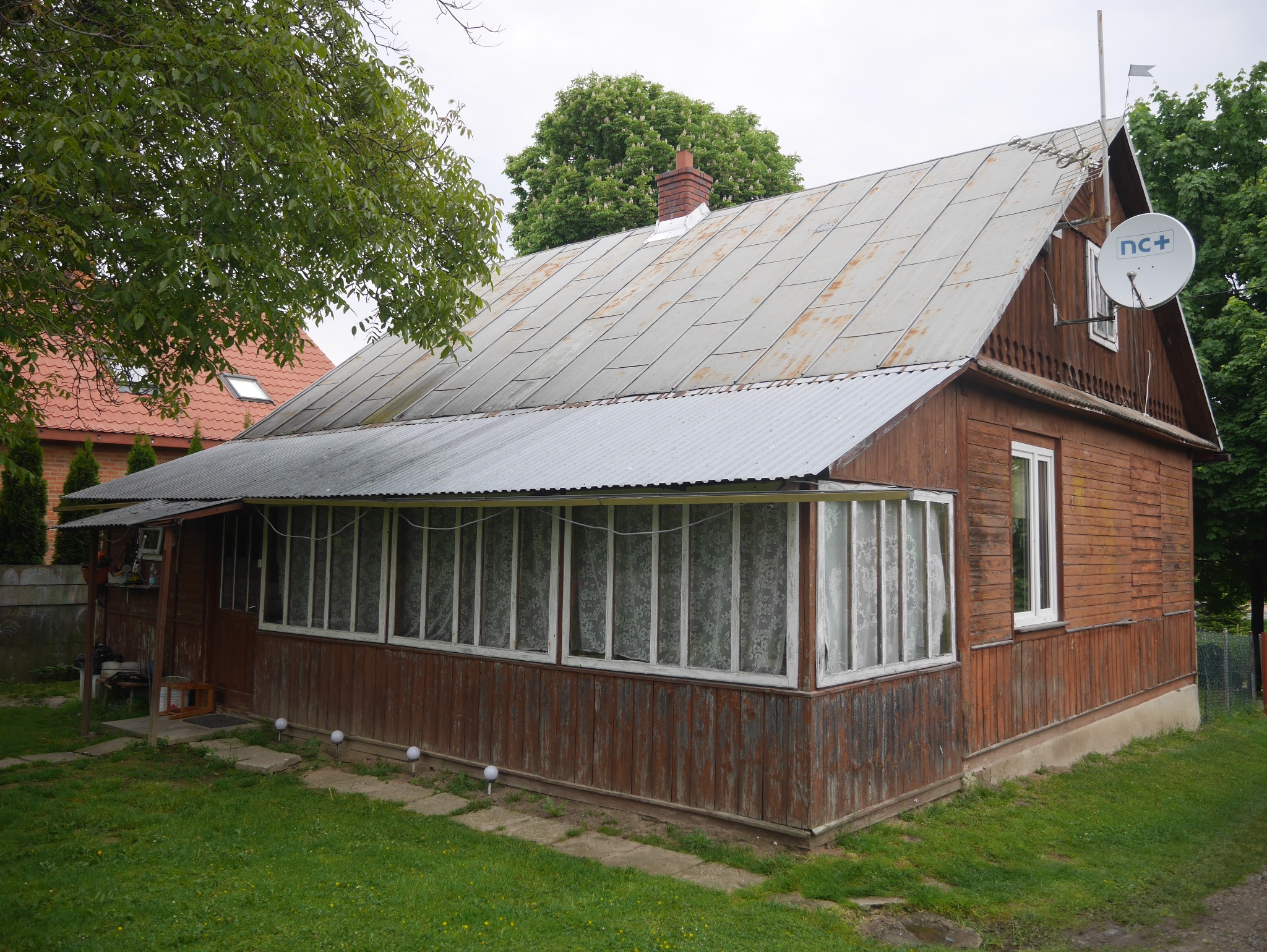 Drewniany dom z blachy falistej na dachu i werandą. Widoczna antena satelitarna. Dom otoczony zielenią. Ujęcie z poziomu trawnika.
