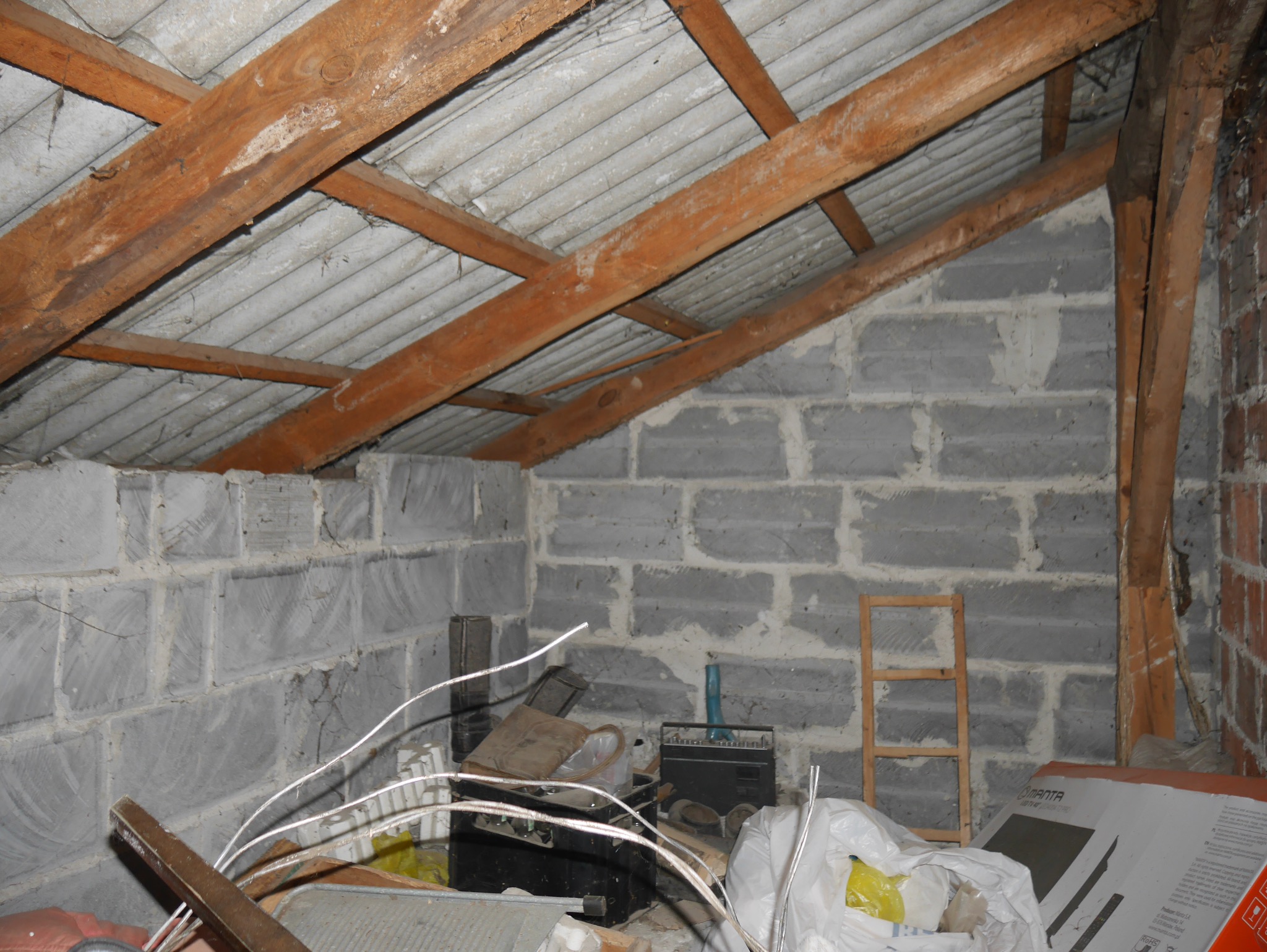 Surowe poddasze z drewnianą konstrukcją dachu i ścianami z szarych bloczków. Widoczne elementy instalacji i akcesoria budowlane. Nierówności i niedokończone wykończenie.