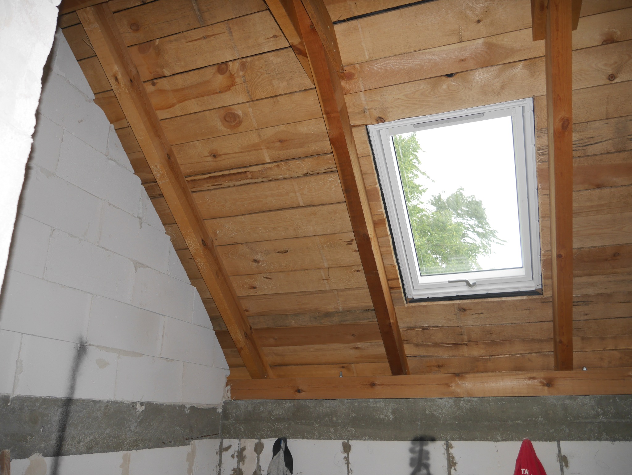 Drewniany strop poddasza z oknem dachowym w trakcie montażu. Widoczne białe bloczki i elementy konstrukcyjne. W tle zieleń za oknem.