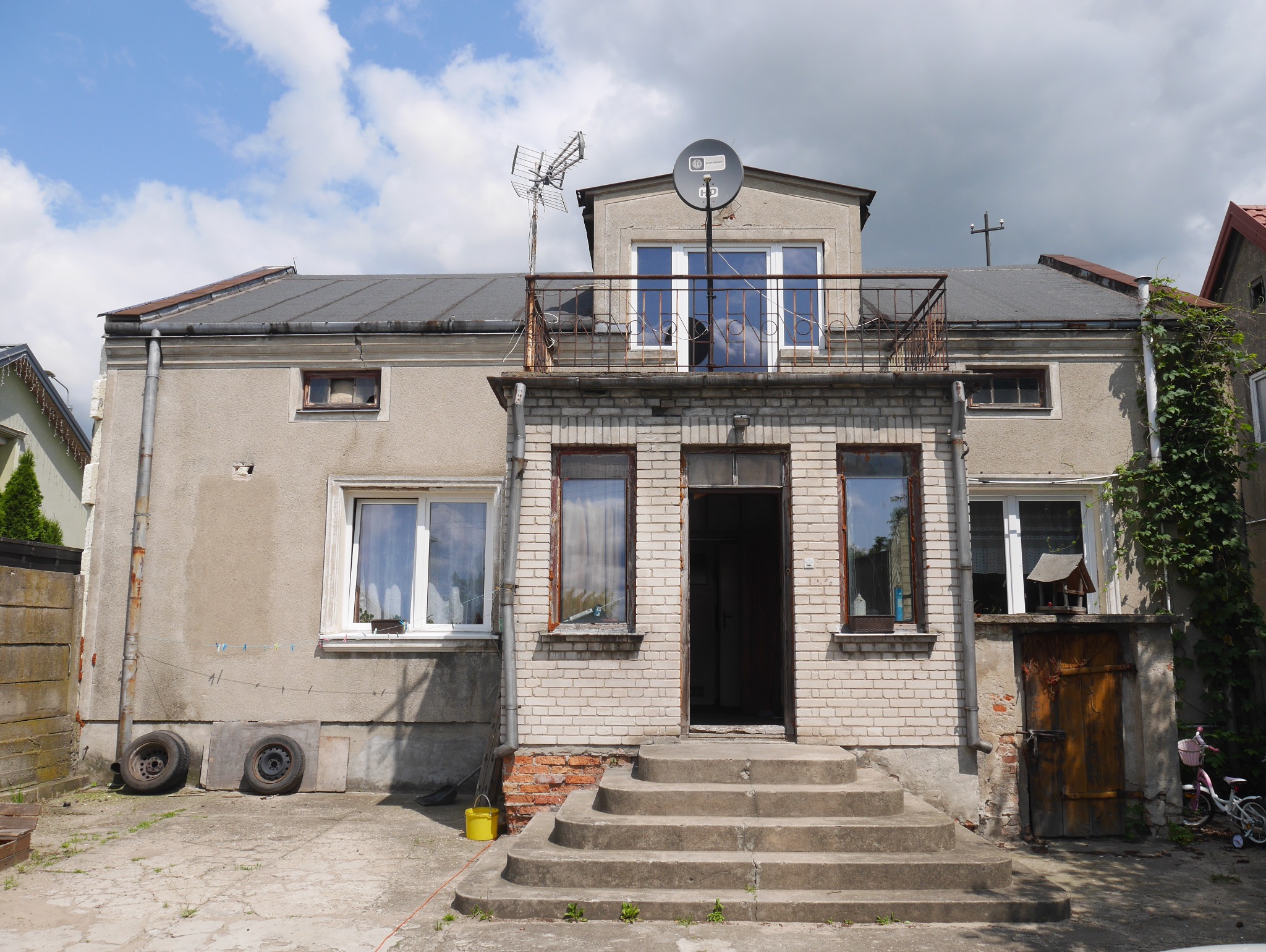 Dwukondygnacyjny dom z ceglaną fasadą, balkonem i schodami wejściowymi. Widoczne okna, drzwi, anteny na dachu i elementy otoczenia, takie jak roślinność i opony.