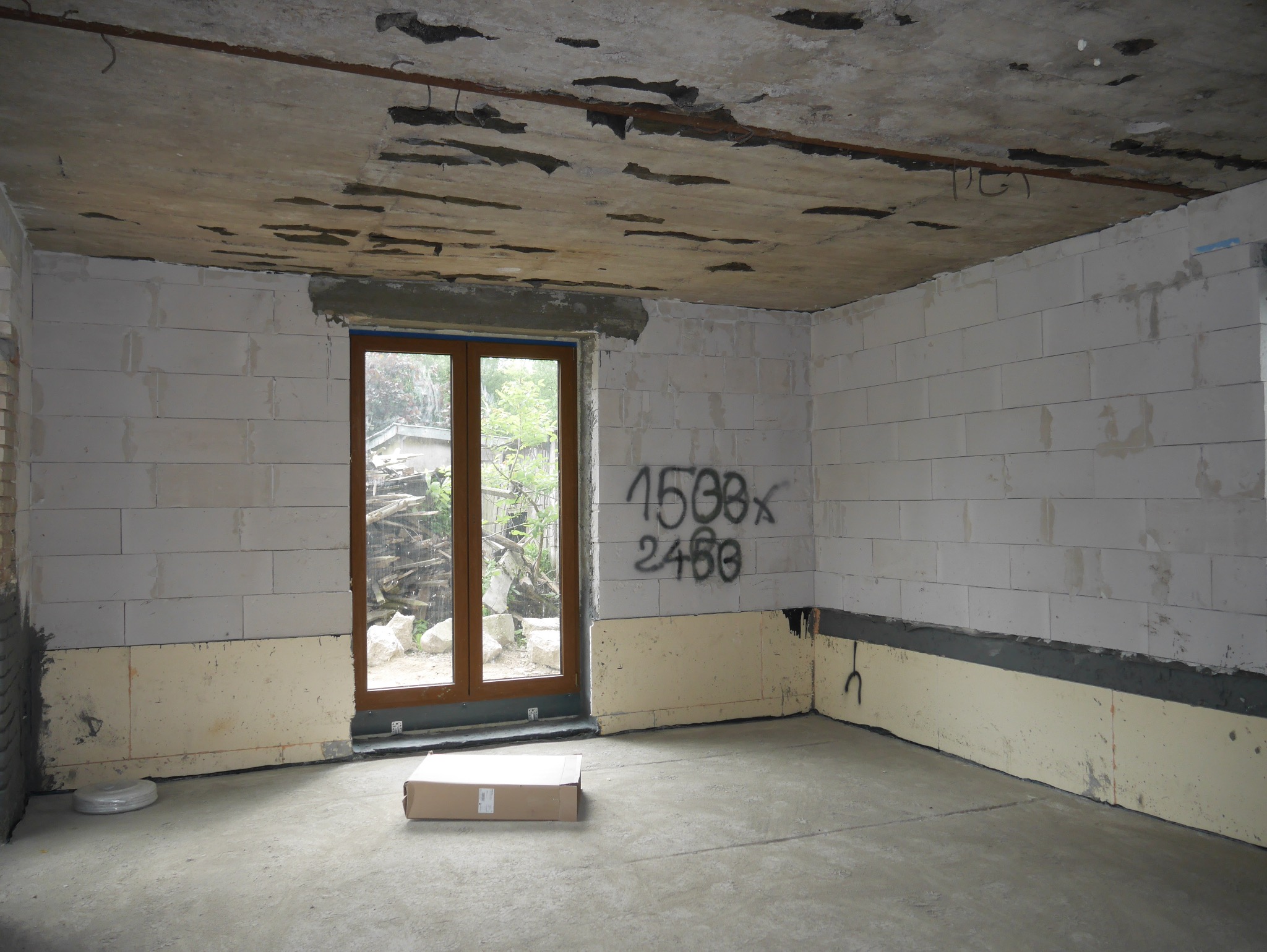 Surowy stan wnętrza: ściany z betonu komórkowego, izolacja, betonowa podłoga. Okno z widokiem na zewnątrz. Na ścianie napisy. Widok na stan deweloperski.