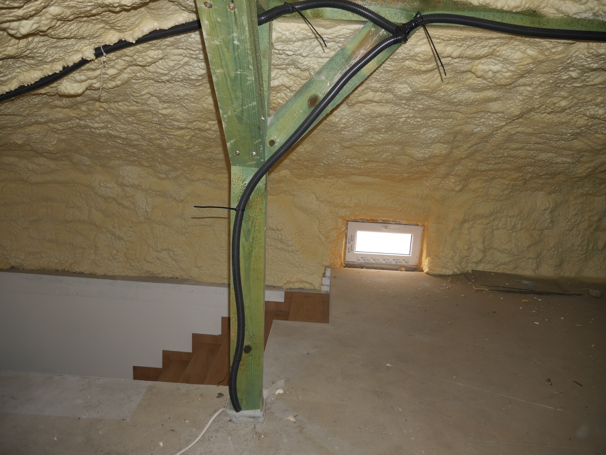 Poddasze po ociepleniu pianą PUR: widoczna gruba warstwa izolacji na skośnym dachu, drewniana konstrukcja wsporcza i instalacja elektryczna. Okno w tle.