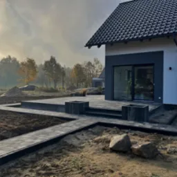 Nowoczesny taras z kostki brukowej w odcieniach szarości, z podwyższonym poziomem i elementami dekoracyjnymi, na tle domu jednorodzinnego w porannej mgle.