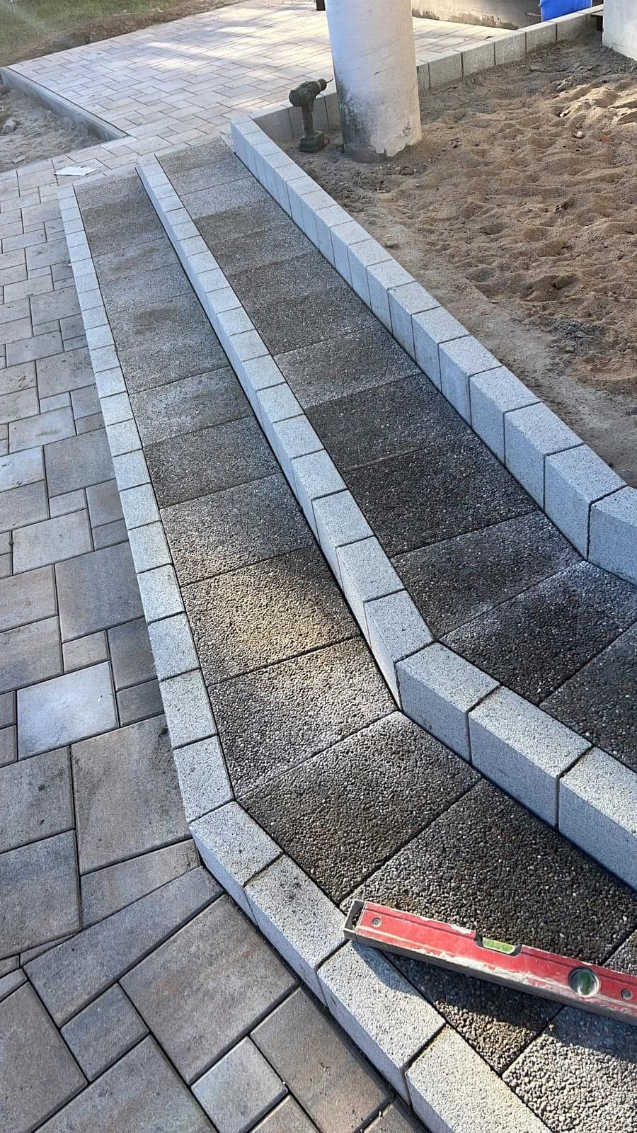 Ułożona kostka brukowa tworząca rampę z szarymi obrzeżami. W tle piasek, wiertarka i poziomica. Widok z góry na fragment chodnika i rampy.