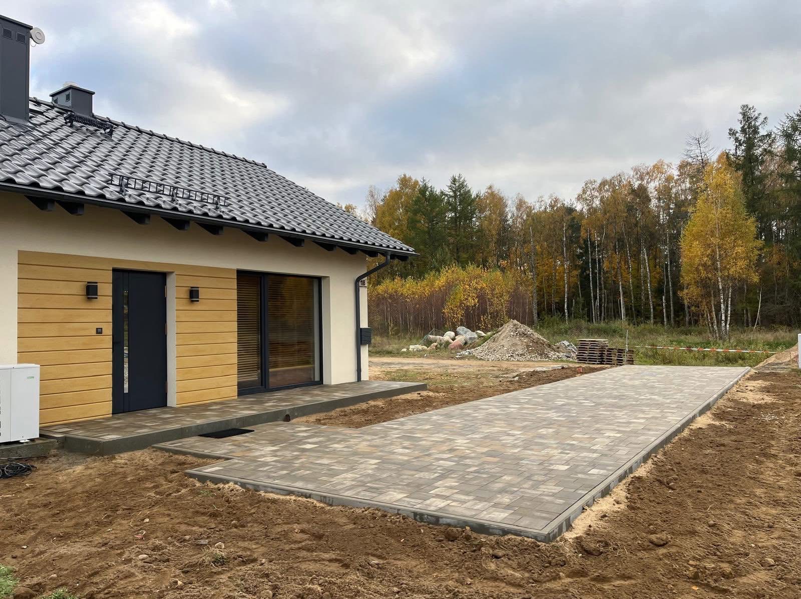 Nowoczesny dom z ułożoną kostką brukową na podjeździe i tarasie. Widoczny fragment elewacji z drewnianym wykończeniem i jesienny las w tle.