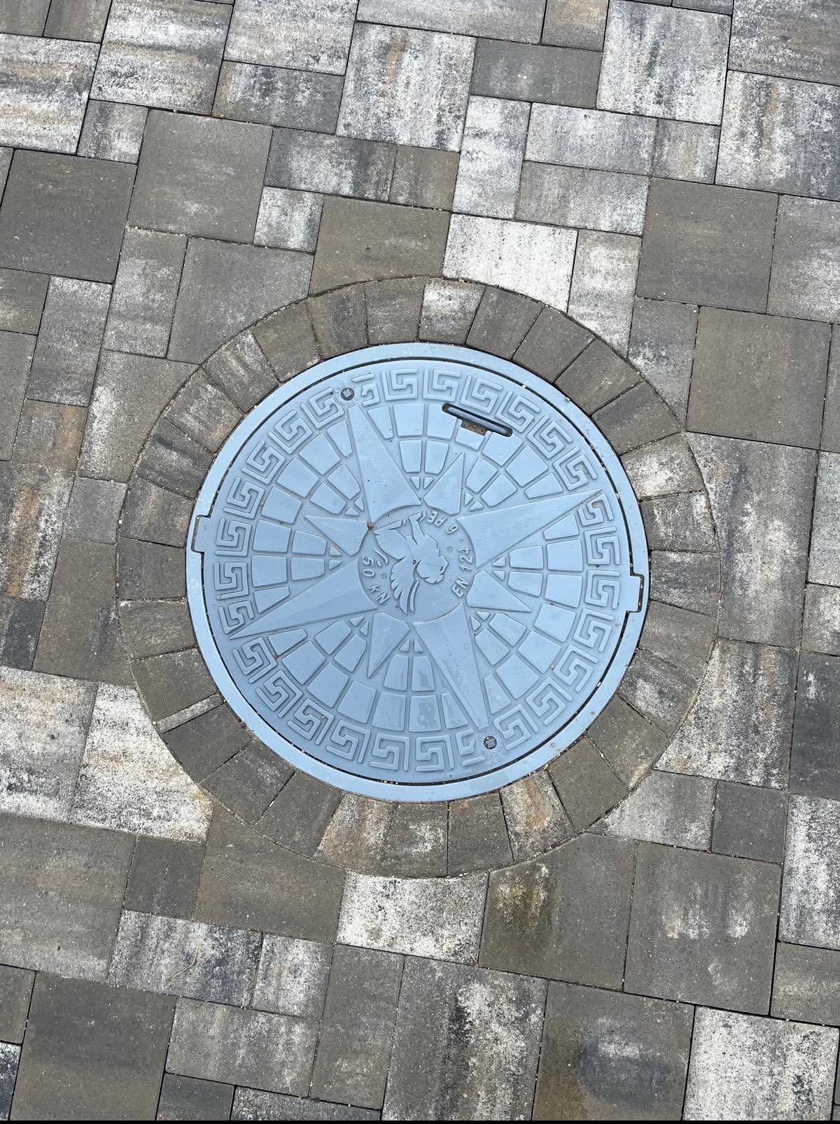 Szara, dekoracyjna studzienka kanalizacyjna z motywem kompasu, wkomponowana w kostkę brukową w Zblewie. Widok z góry, regularny wzór kostki.