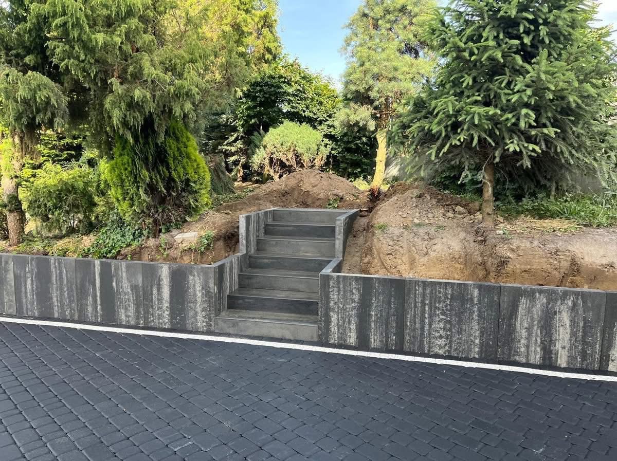Nowoczesne schody z betonowych elementów prowadzące na wzniesienie, otoczone zielenią. Ułożona kostka brukowa i mur oporowy z betonu.
