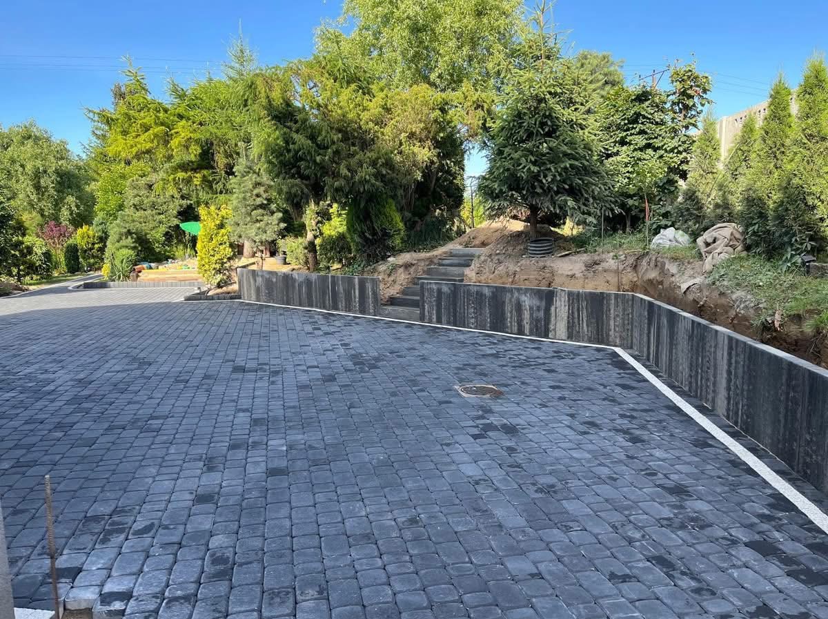 Wykończony podjazd z kostki brukowej w odcieniach szarości, otoczony zielenią. Widoczna mur oporowy i schody prowadzące do ogrodu.