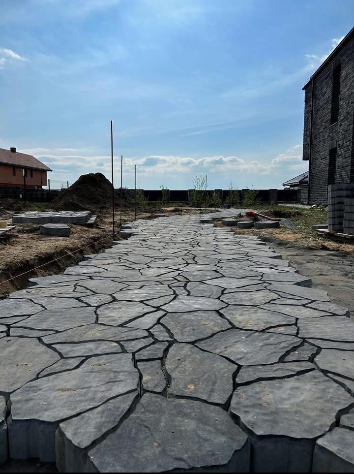 Ścieżka z nieregularnych, szarych płyt kamiennych w trakcie układania, z widocznym przygotowaniem terenu i stosem płyt w tle, dom w tle.