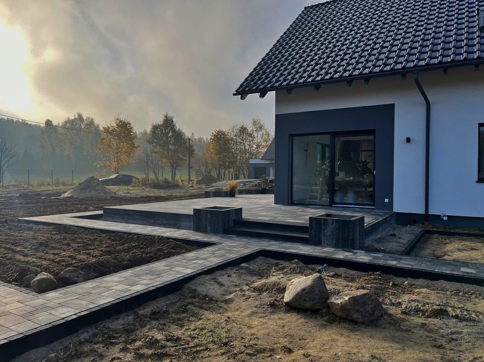 Nowoczesny taras z kostki brukowej w odcieniach szarości, z podwyższonym poziomem i elementami dekoracyjnymi, na tle domu jednorodzinnego w porannej mgle.