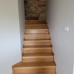 Jasne, dębowe schody z delikatnym usłojeniem, prowadzące w górę, z widoczną ścianą z cegły w tle. Podłoga wykończona parkietem w jodełkę.