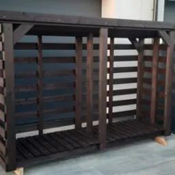 Ciemnobrązowa, drewniana konstrukcja na drewno, z poziomymi szczebelkami, ustawiona na kostce brukowej, na małych klinach. Tło: budynek i ogrodzenie.