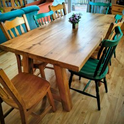 TRIMA Szymon Zielonka - Solidny, dębowy stół z kwiatami w centralnej części, otoczony różnokolorowymi krzesłami. Widoczne naturalne usłojenie drewna i masywna konstrukcja stołu.