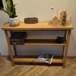 TRIMA Szymon Zielonka - Drewniany stolik konsola z dwoma półkami, na blacie dekoracje: roślina w doniczce z twarzą, koszyk, świece i drewniane ozdoby. Na półkach książki i aparat.