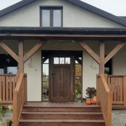 TRIMA Szymon Zielonka - Wejście do domu z drewnianymi schodami i balustradą, zadaszone drewnianą konstrukcją. Widoczne dynie i rośliny w donicach. Dom z oknem na poddaszu.