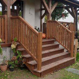 TRIMA Szymon Zielonka - Drewniane schody wejściowe z balustradą, prowadzące do zadaszonego tarasu. Udekorowane dynią i kwiatami w donicach. Widoczne elementy konstrukcji drewnianej.