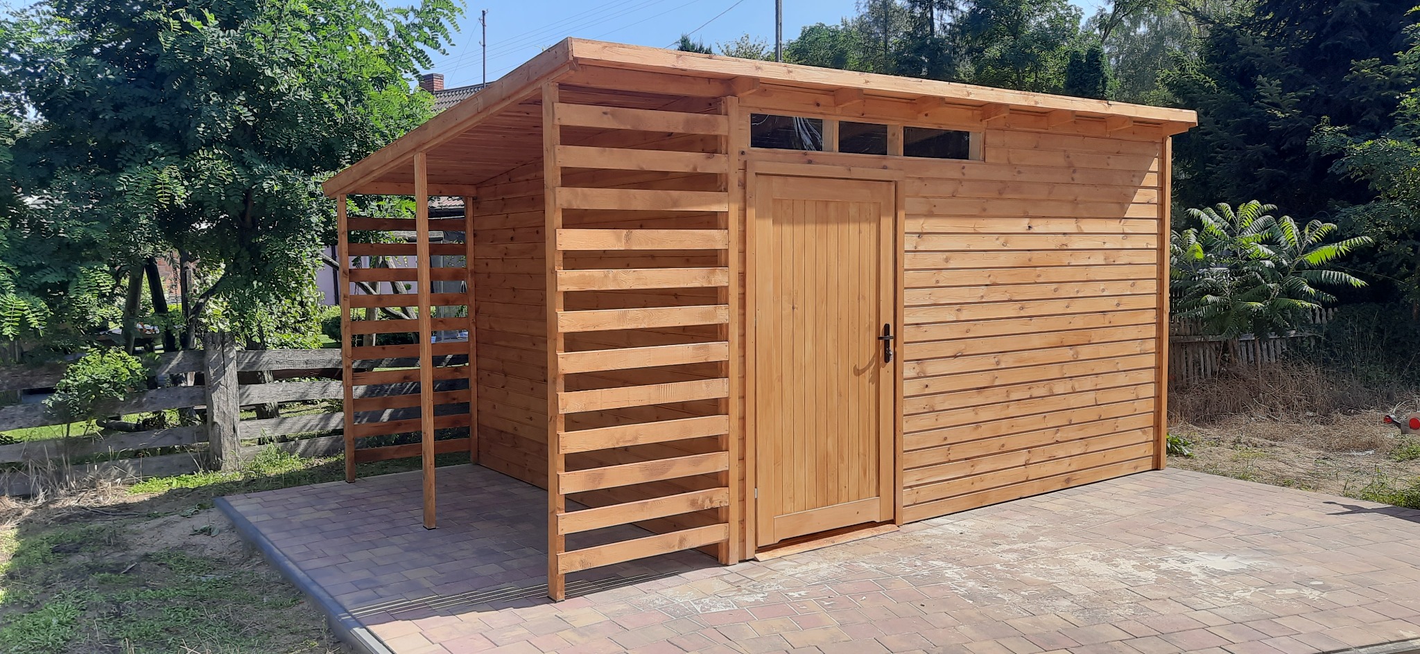 Drewniana altana z zadaszeniem i ażurowymi ścianami, usytuowana na kostce brukowej, otoczona zielenią. Naturalne oświetlenie podkreśla strukturę drewna.