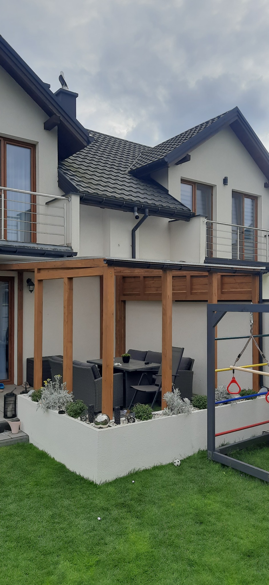 Pergola z drewnianą konstrukcją i zadaszeniem, przylegająca do ściany domu, z meblami ogrodowymi i dekoracyjną rabatą. Obok plac zabaw.