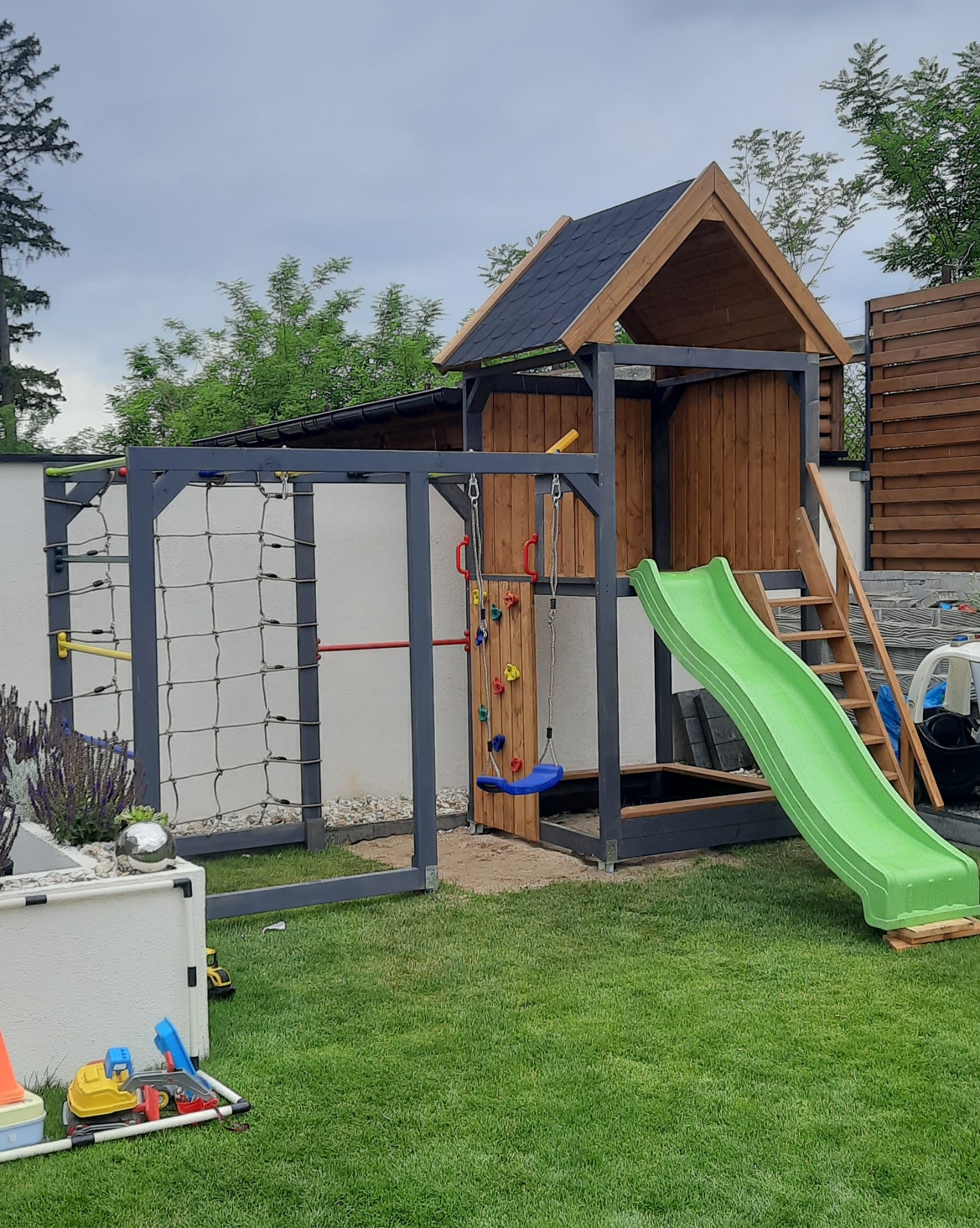 Drewniany plac zabaw z domkiem, ścianką wspinaczkową, siatką i zjeżdżalnią na trawniku. Konstrukcja pomalowana na szaro, zadaszenie z ciemnej dachówki.