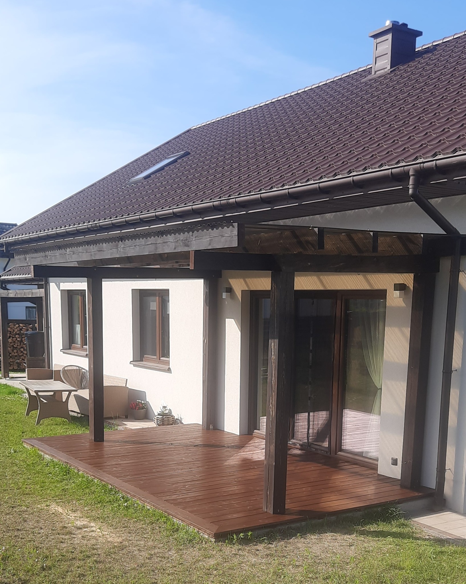 Drewniany taras przy domu z brązowymi belkami i podłogą, widok na okna i drzwi tarasowe. W tle meble ogrodowe i zielony trawnik.
