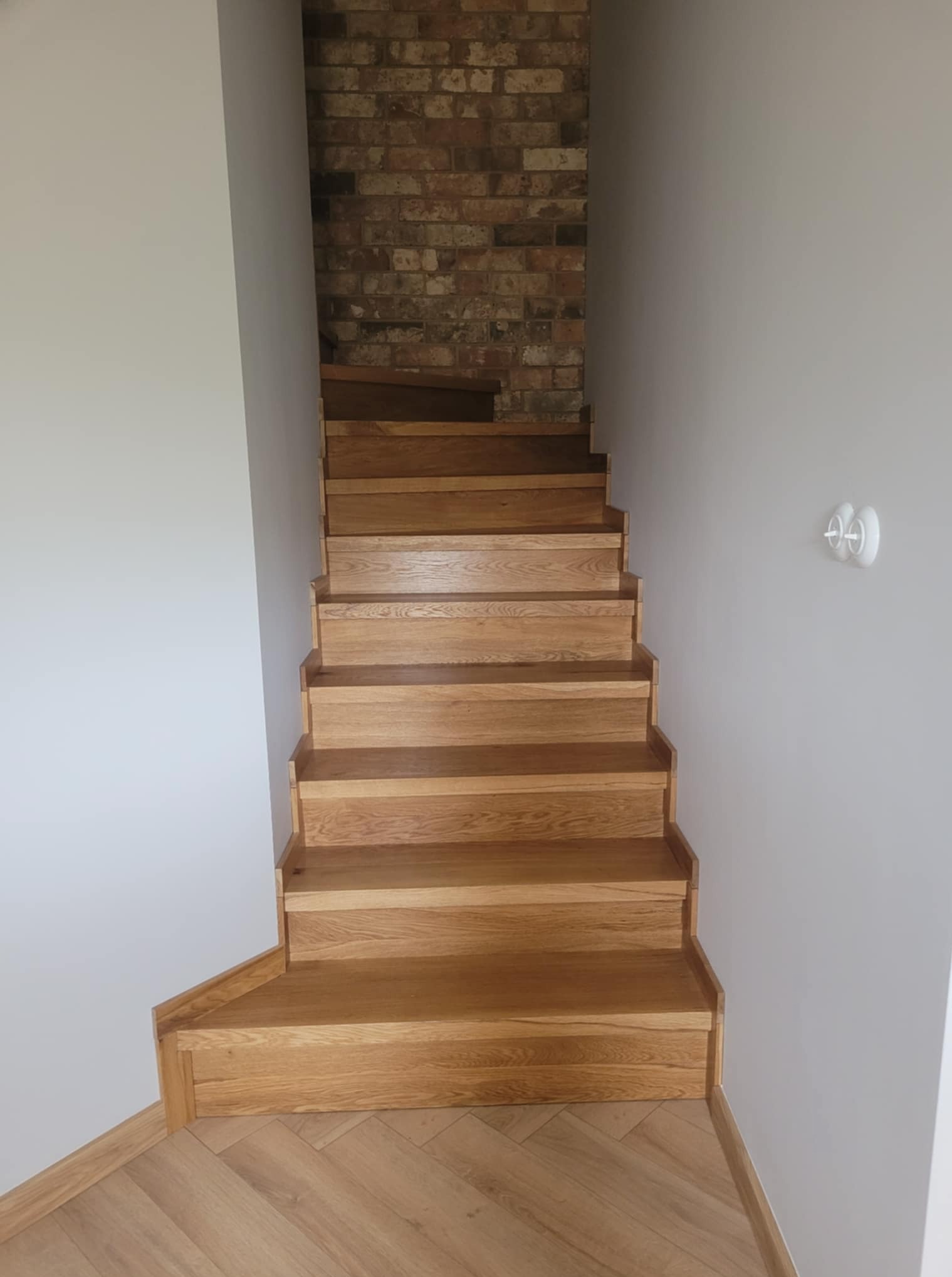 Jasne, dębowe schody z delikatnym usłojeniem, prowadzące w górę, z widoczną ścianą z cegły w tle. Podłoga wykończona parkietem w jodełkę.