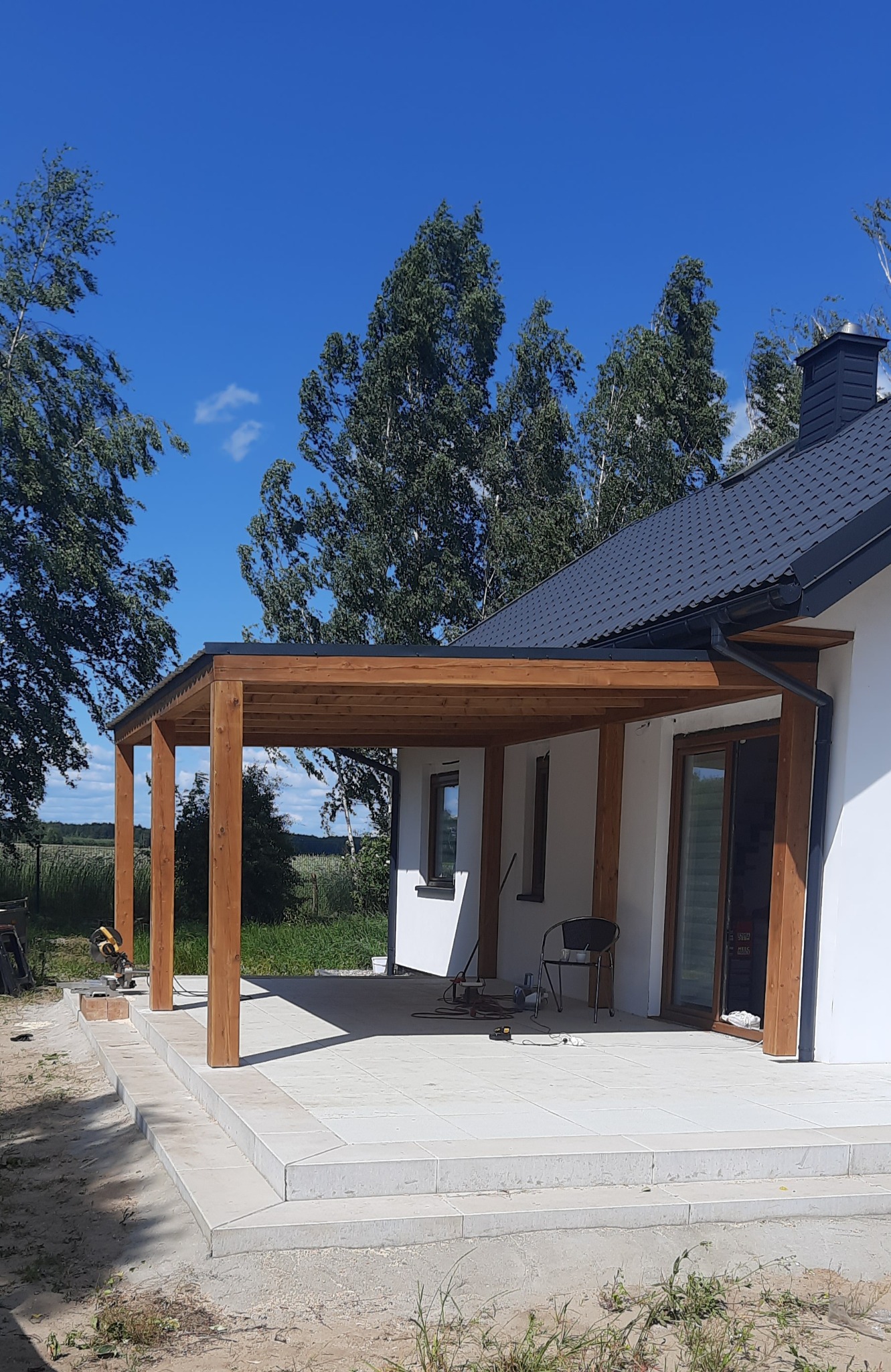 Drewniana konstrukcja zadaszenia tarasu przy białym domu z czarnym dachem, widok na jasną, wyłożoną płytkami podłogę i zieleń w tle, słoneczny dzień.