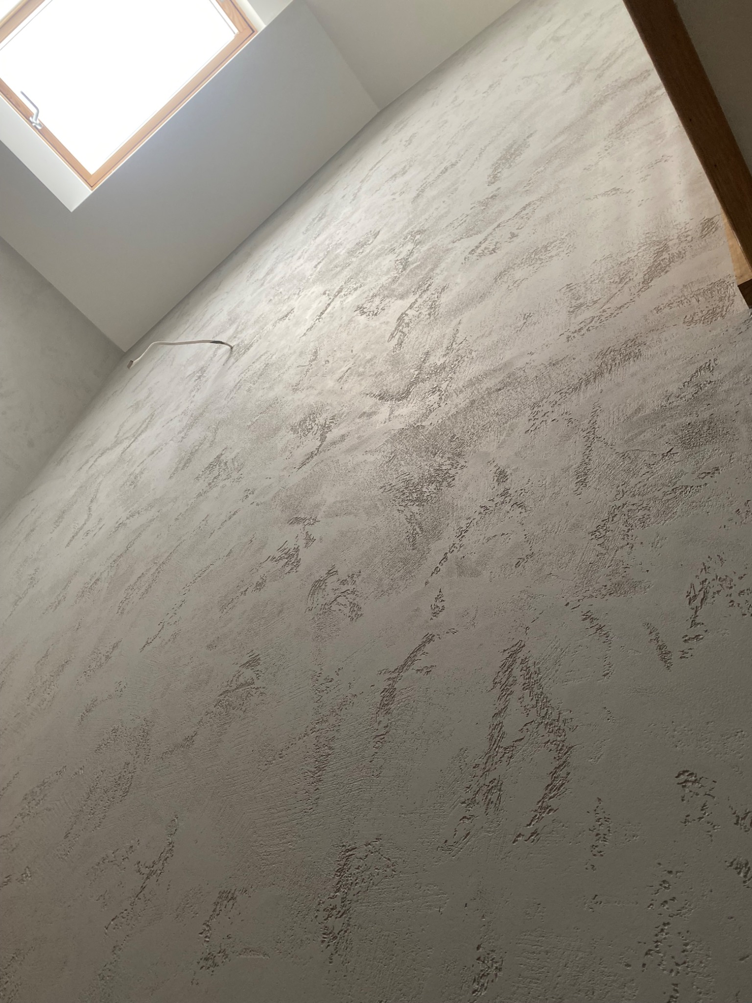 Ściana z dekoracyjną fakturą w odcieniach szarości, oświetlona światłem z okna dachowego. Widoczny fragment drewnianej ramy okiennej i przewód elektryczny.