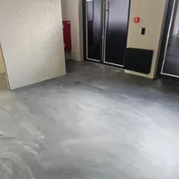 Błyszcząca, szara posadzka w pomieszczeniu z grzejnikiem i drzwiami, imitująca wzorem marmurkowe żyły; widoczna instalacja przeciwpożarowa.