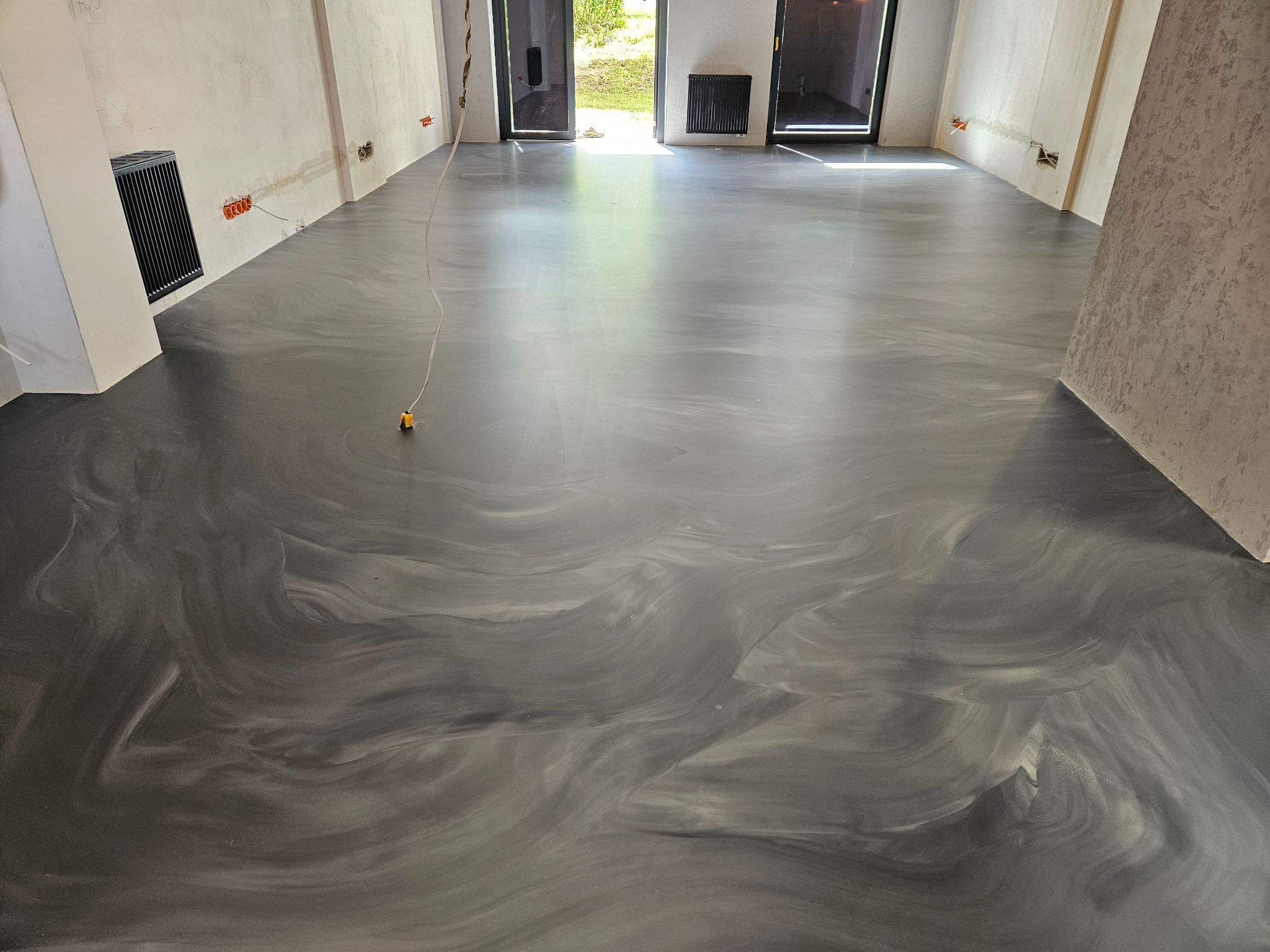 Błyszcząca, szara posadzka żywiczna z unikalnym wzorem przypominającym marmur w surowym wnętrzu z widocznymi instalacjami elektrycznymi i grzejnikami ściennymi.