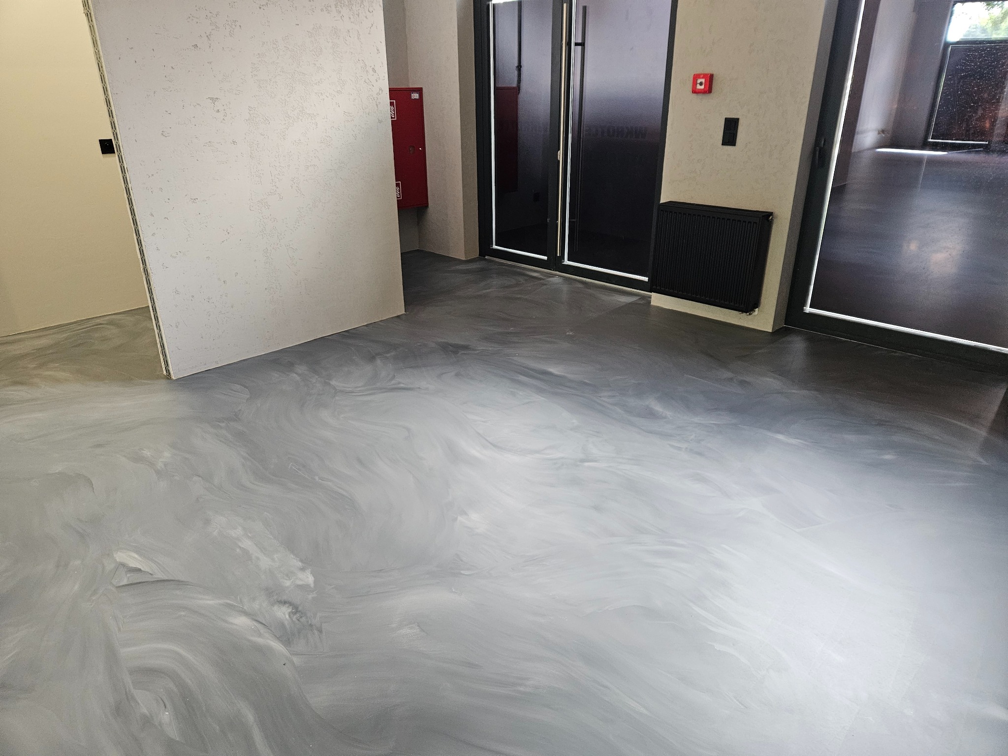 Błyszcząca, szara posadzka w pomieszczeniu z grzejnikiem i drzwiami, imitująca wzorem marmurkowe żyły; widoczna instalacja przeciwpożarowa.