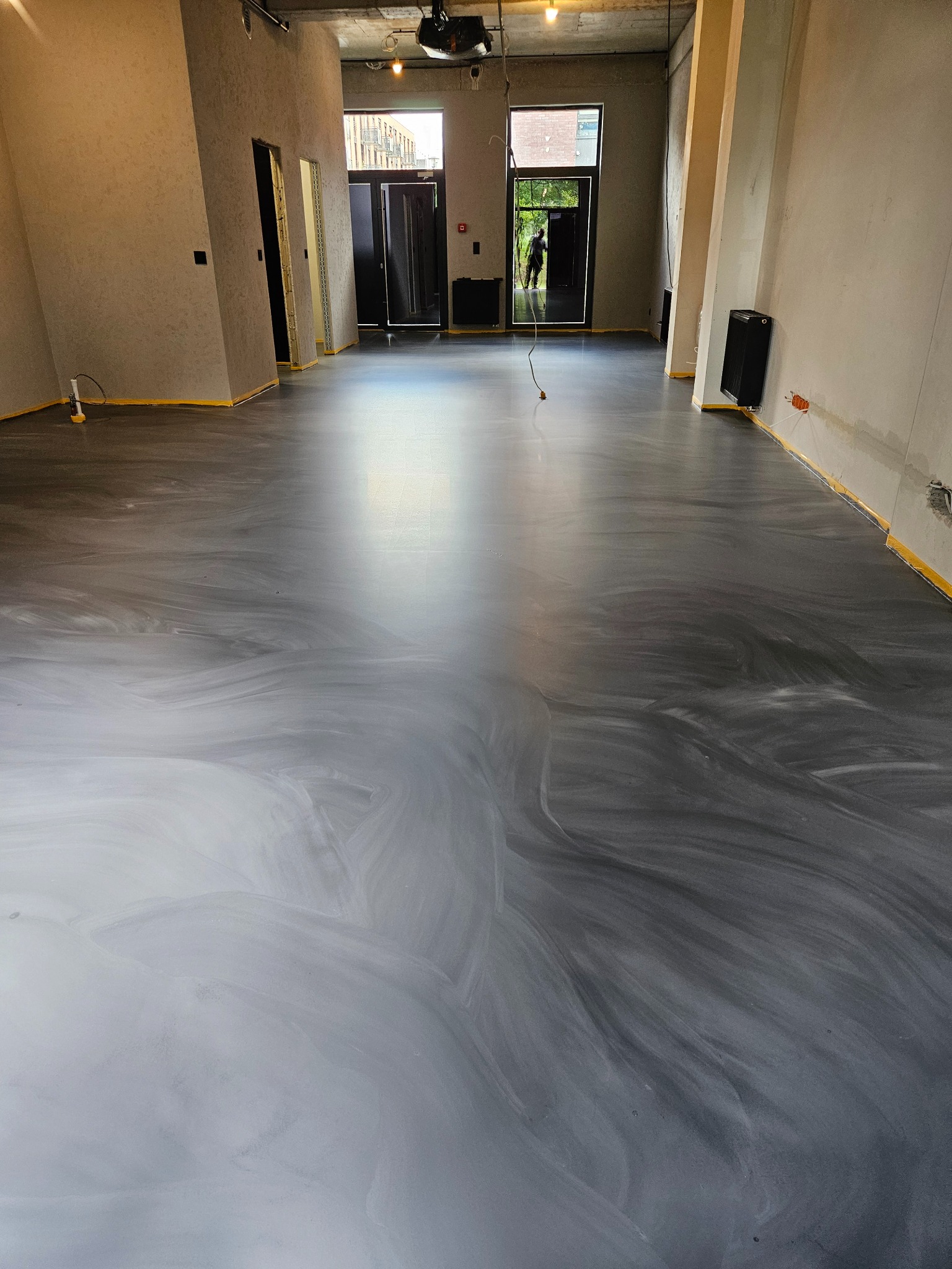 Szara, błyszcząca posadzka w przestronnym wnętrzu z surowymi ścianami. Widoczne taśmy zabezpieczające przy ścianach i osoba w drzwiach na końcu pomieszczenia.