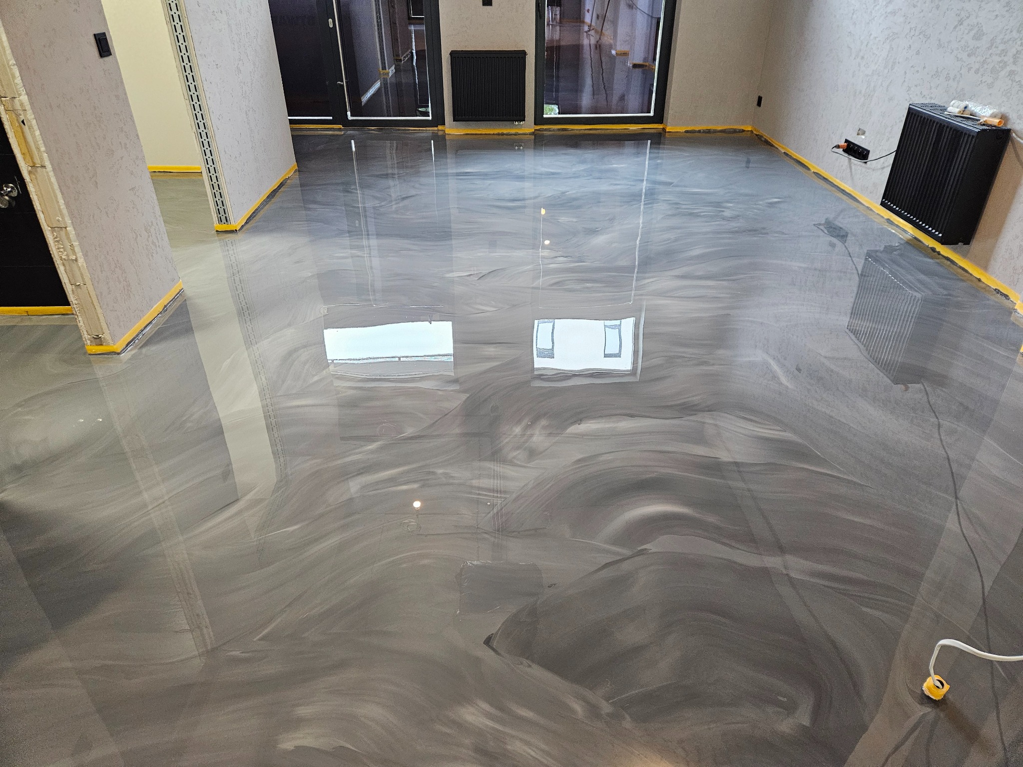 Błyszcząca, szara posadzka z efektem marmuru w przestronnym wnętrzu. Żółta taśma zabezpieczająca przy ścianach. Widoczne odbicie okien w gładkiej powierzchni.