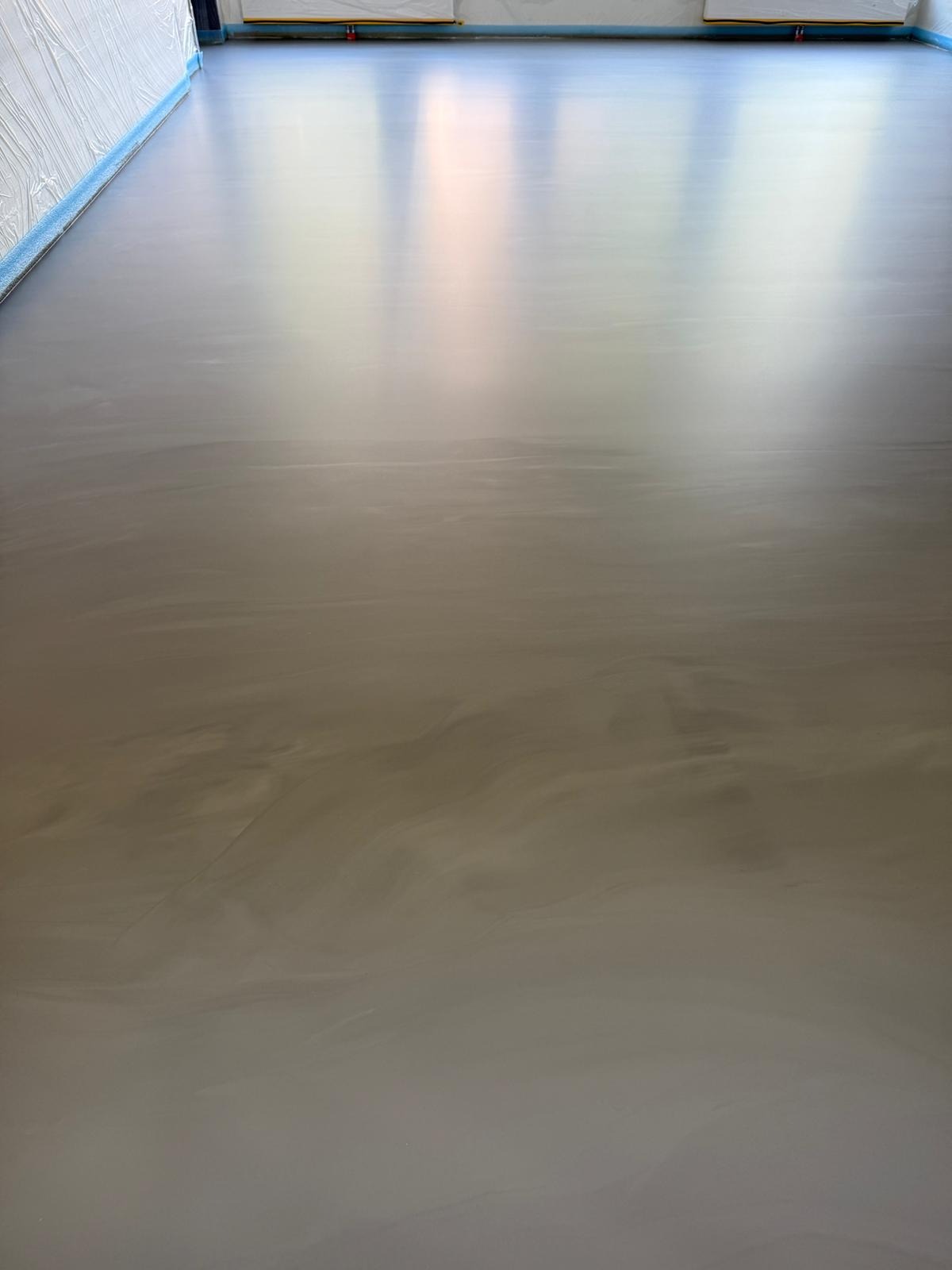 Błyszcząca, beżowo-szara posadzka z efektem chmur, świeżo wylana, zabezpieczona folią przy ścianach. Widoczne odbicia światła.