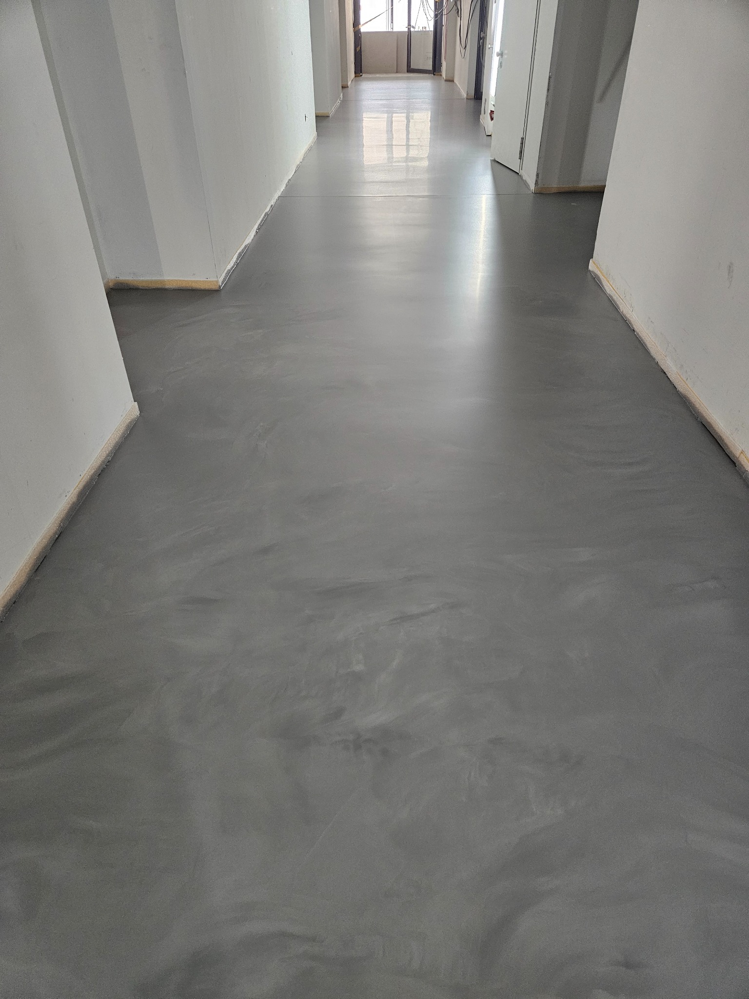 Szara, gładka posadzka w długim korytarzu z białymi ścianami, widok z perspektywy, delikatne refleksy światła na powierzchni podłogi.