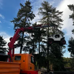 Pomarańczowy dźwig podnosi biały samochód terenowy Suzuki na tle drzew i błękitnego nieba z chmurami. Widoczny fragment skrzyni ładunkowej ciężarówki.
