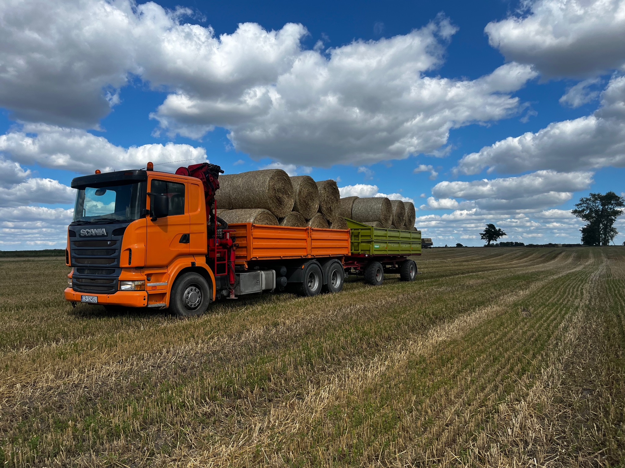 Pomarańczowa ciężarówka Scania z przyczepą, załadowana balotami siana, na tle pola po żniwach i błękitnego nieba z chmurami.