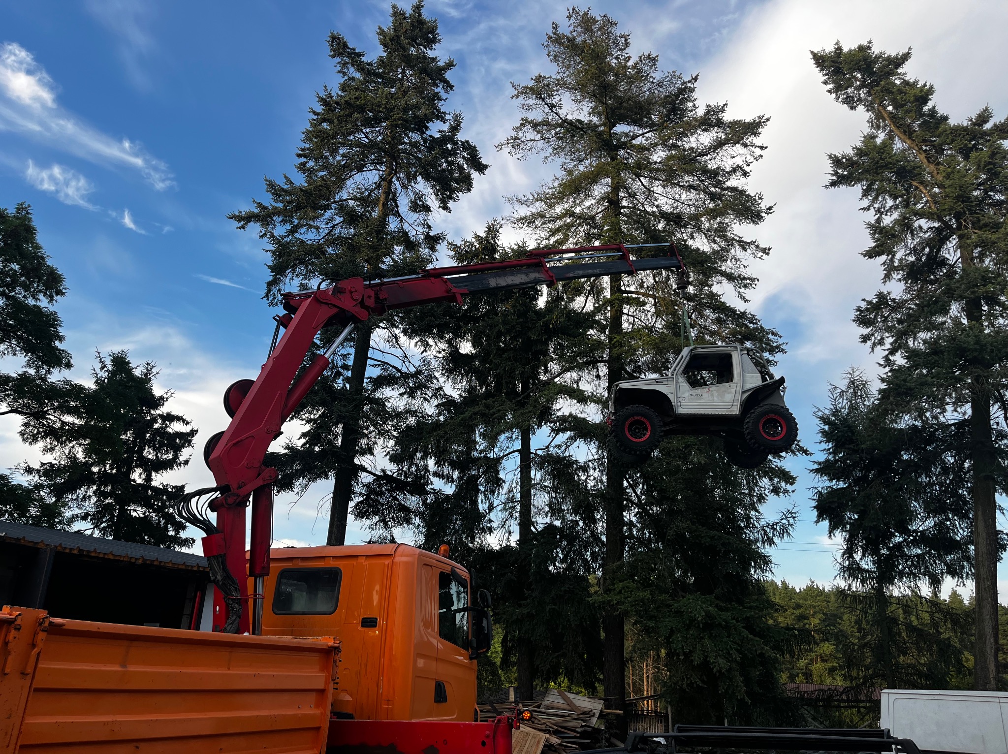 Pomarańczowy dźwig podnosi biały samochód terenowy Suzuki na tle drzew i błękitnego nieba z chmurami. Widoczny fragment skrzyni ładunkowej ciężarówki.