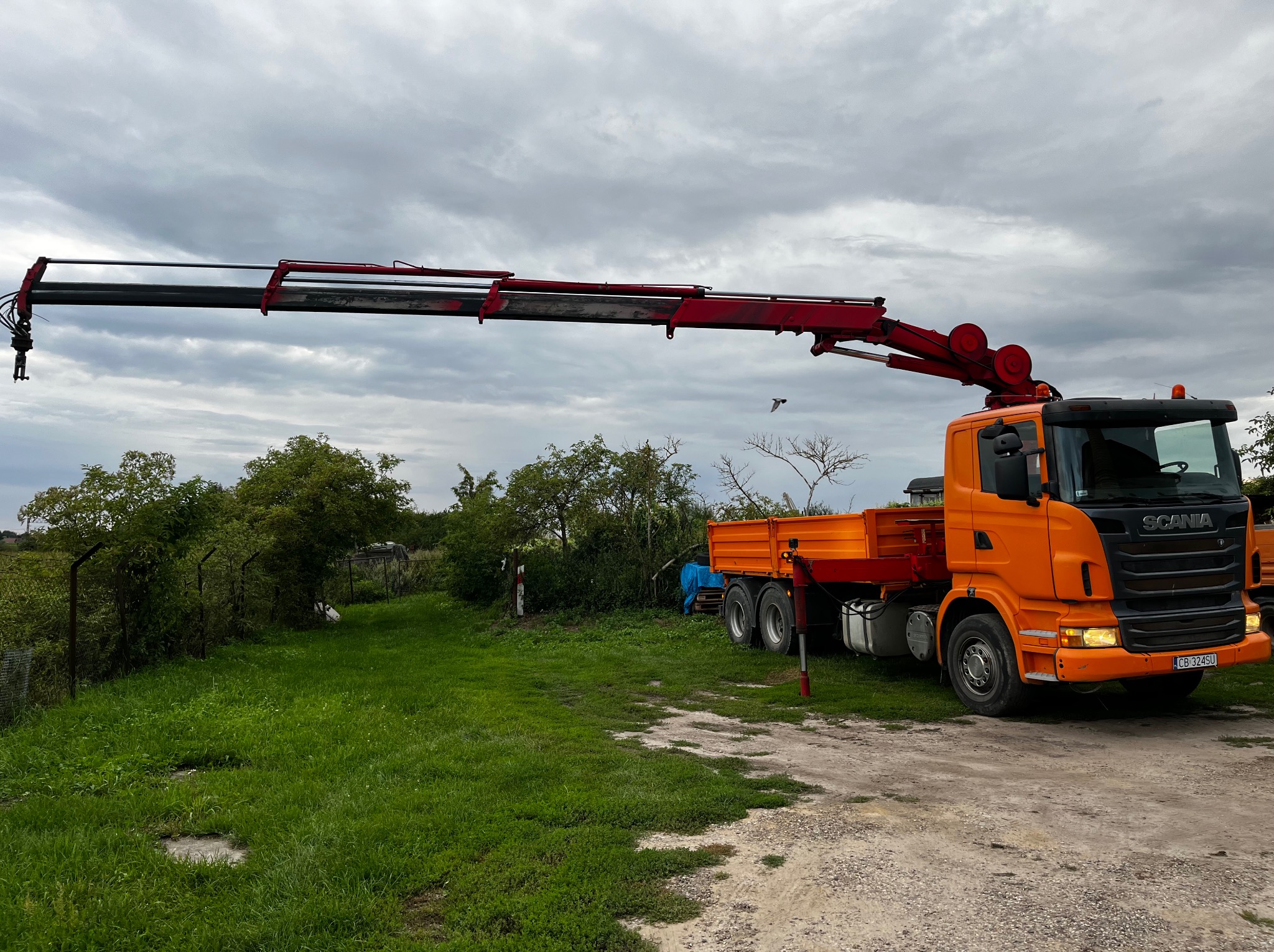 Pomarańczowa ciężarówka Scania z wysięgnikiem dźwigowym na tle zachmurzonego nieba i zieleni. Widoczny numer rejestracyjny pojazdu. Dźwig gotowy do pracy.