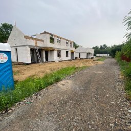 Vitalii Vitalii - Budowa domu z białych bloczków, widoczne ściany i otwory okienne. Na pierwszym planie toaleta przenośna, w tle kolejne budynki w stanie surowym otwartym.