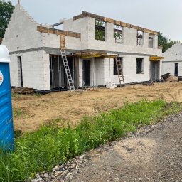 Vitalii Vitalii - Budowa domu z białych bloczków, widoczne rusztowania i drabiny, teren budowy z zielenią. Na pierwszym planie przenośna toaleta.