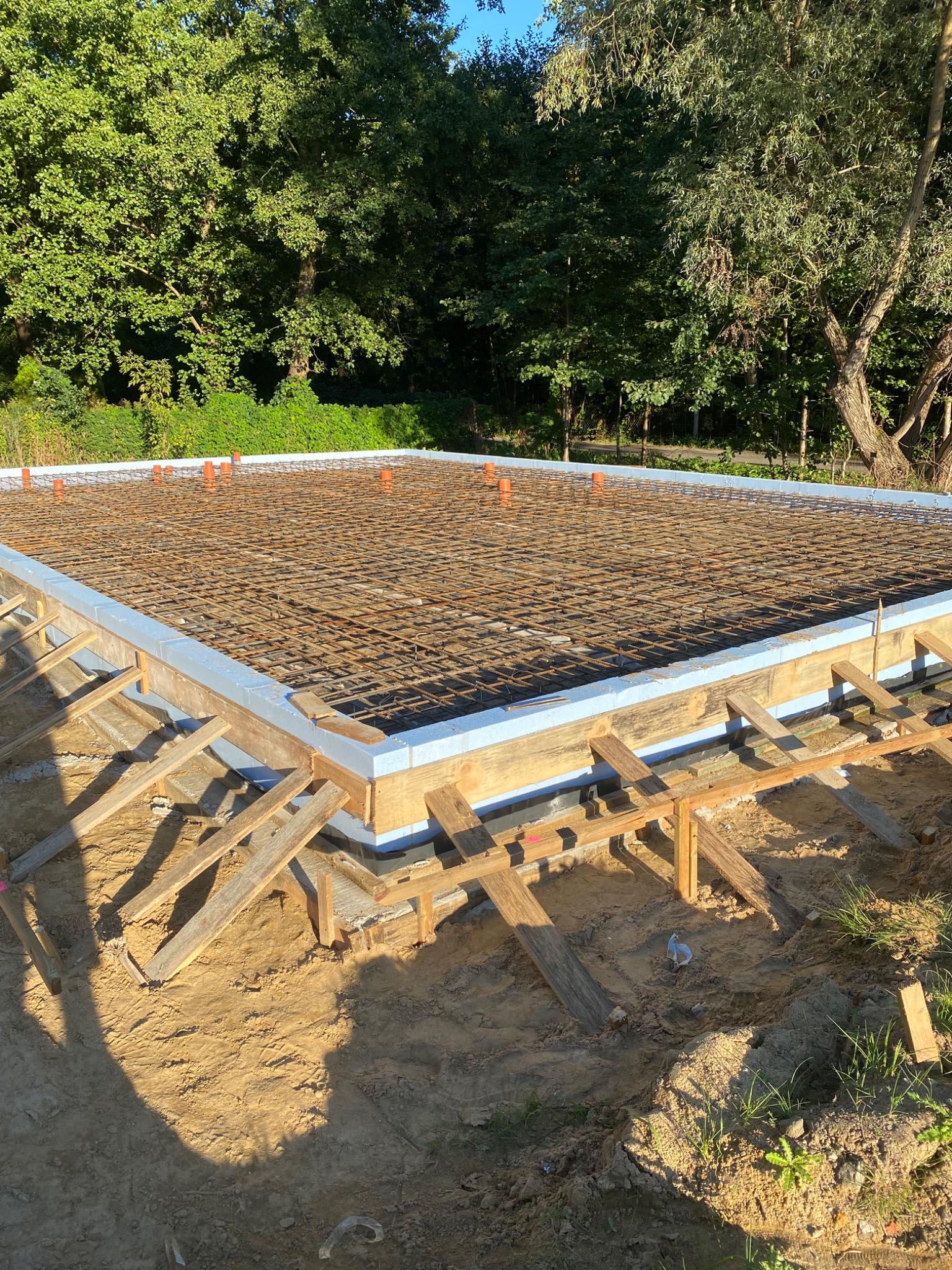 Przygotowany fundament z deskowaniem i zbrojeniem na tle zieleni. Widoczne pomarańczowe rury i drewniane podpory. Kąt ujęcia z góry.