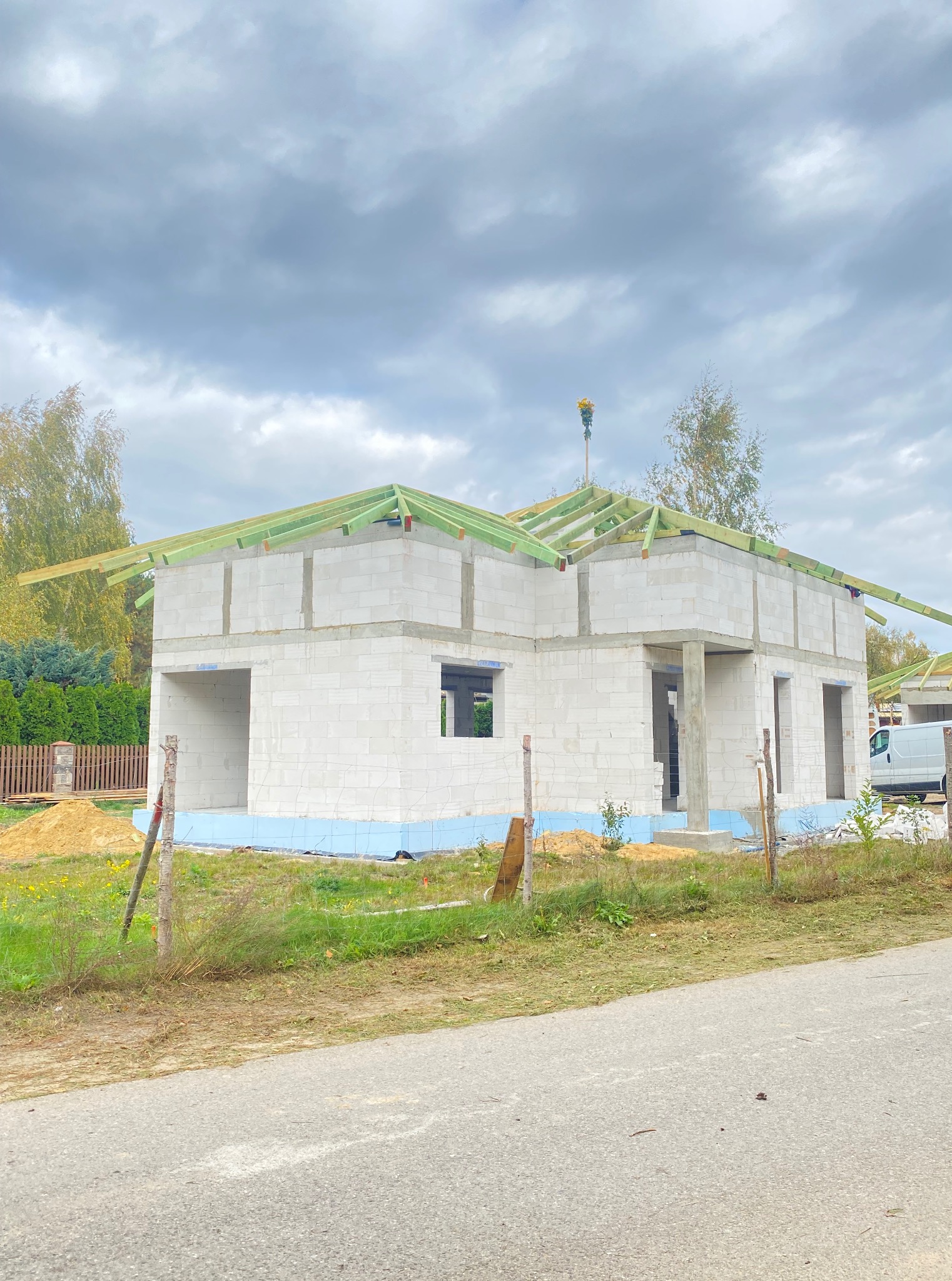 Wznoszenie parterowego domu z betonu komórkowego. Widoczna niebieska izolacja fundamentów i konstrukcja dachu z zielonych belek. W tle pochmurne niebo.