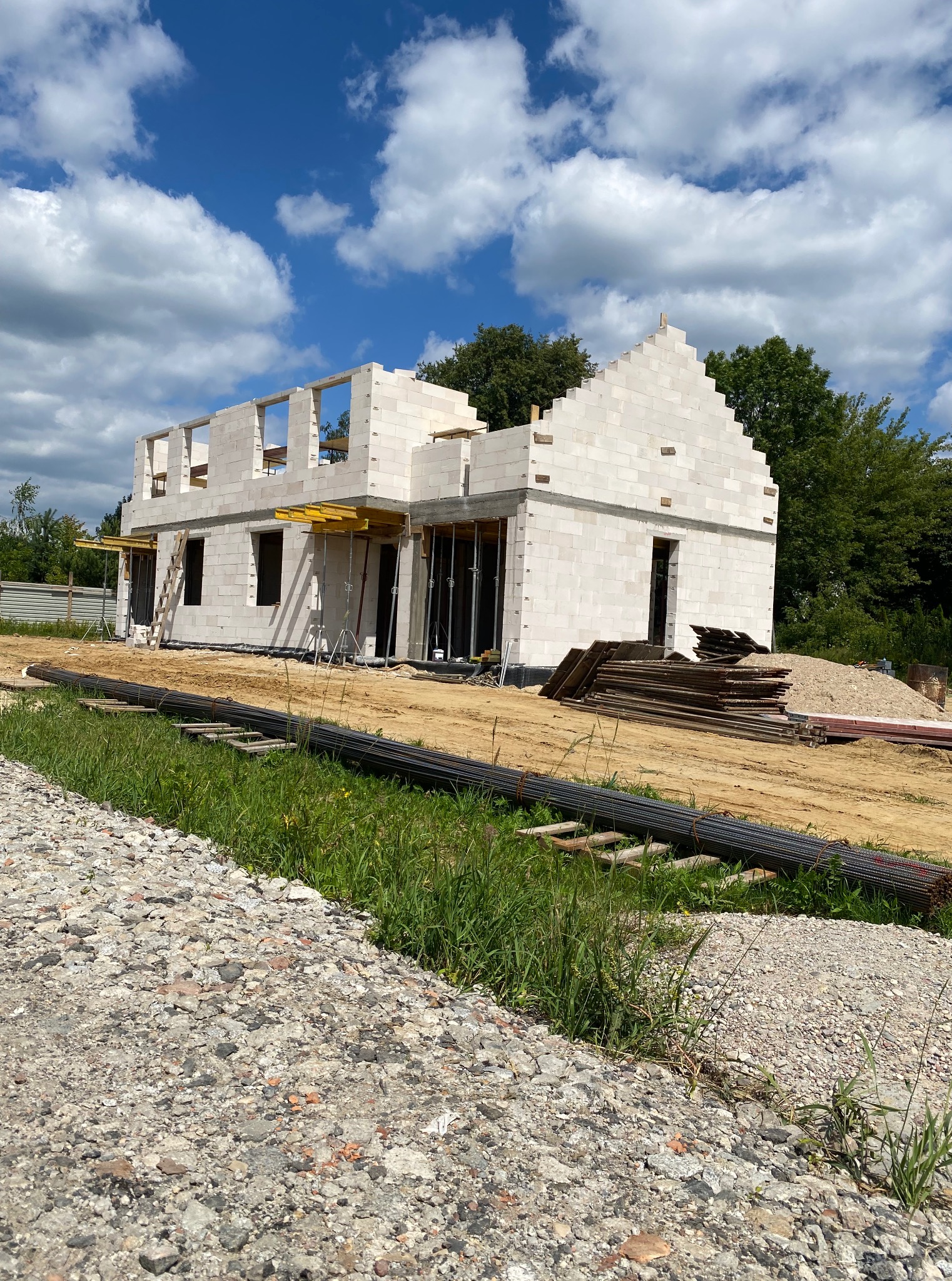 Budowa domu z białych bloczków: dwukondygnacyjny budynek w stanie surowym otwartym, widoczne zbrojenia, drewniane stemple, błękitne niebo z chmurami w tle.