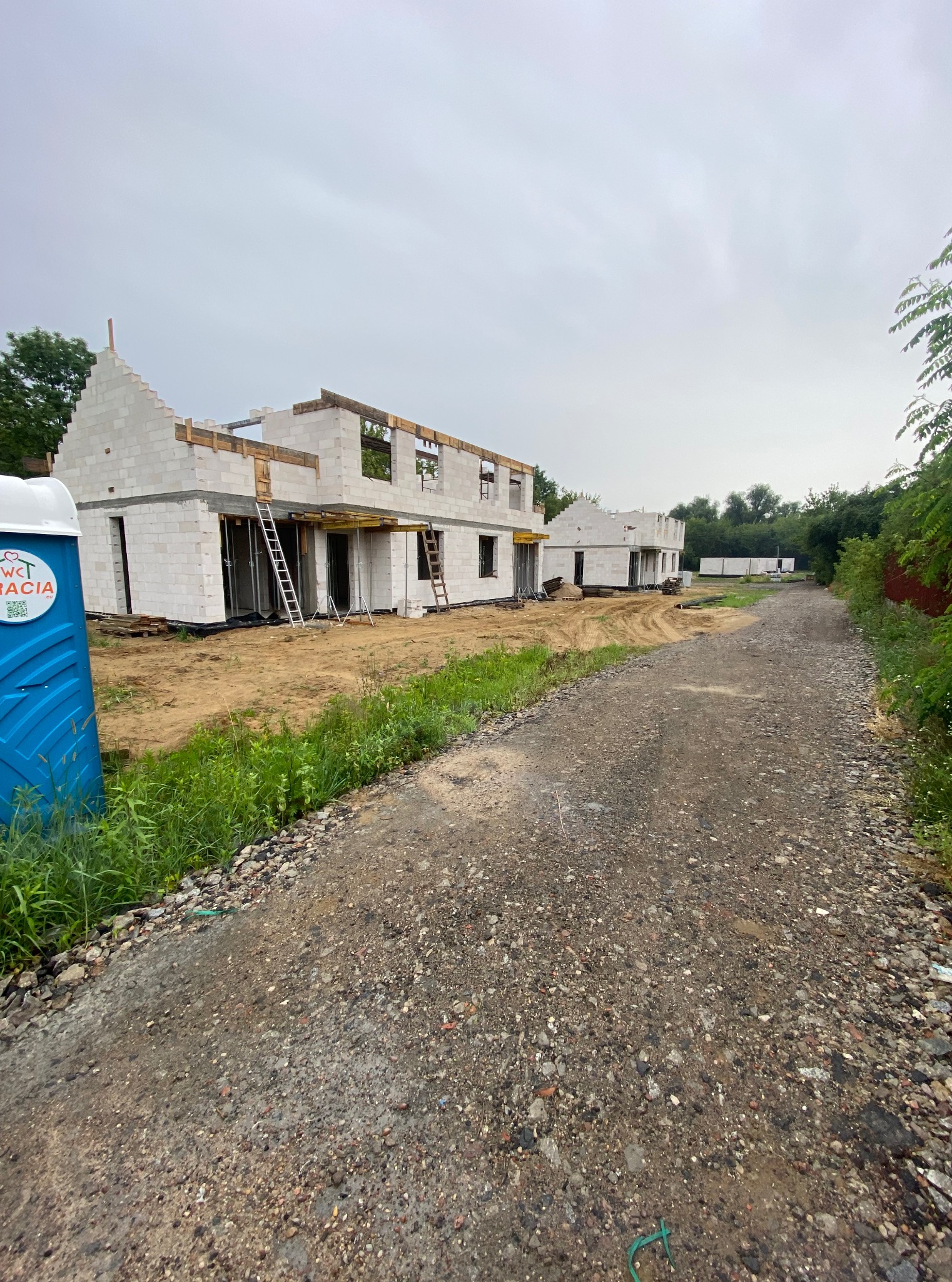 Budowa domu z białych bloczków, widoczne ściany i otwory okienne. Na pierwszym planie toaleta przenośna, w tle kolejne budynki w stanie surowym otwartym.