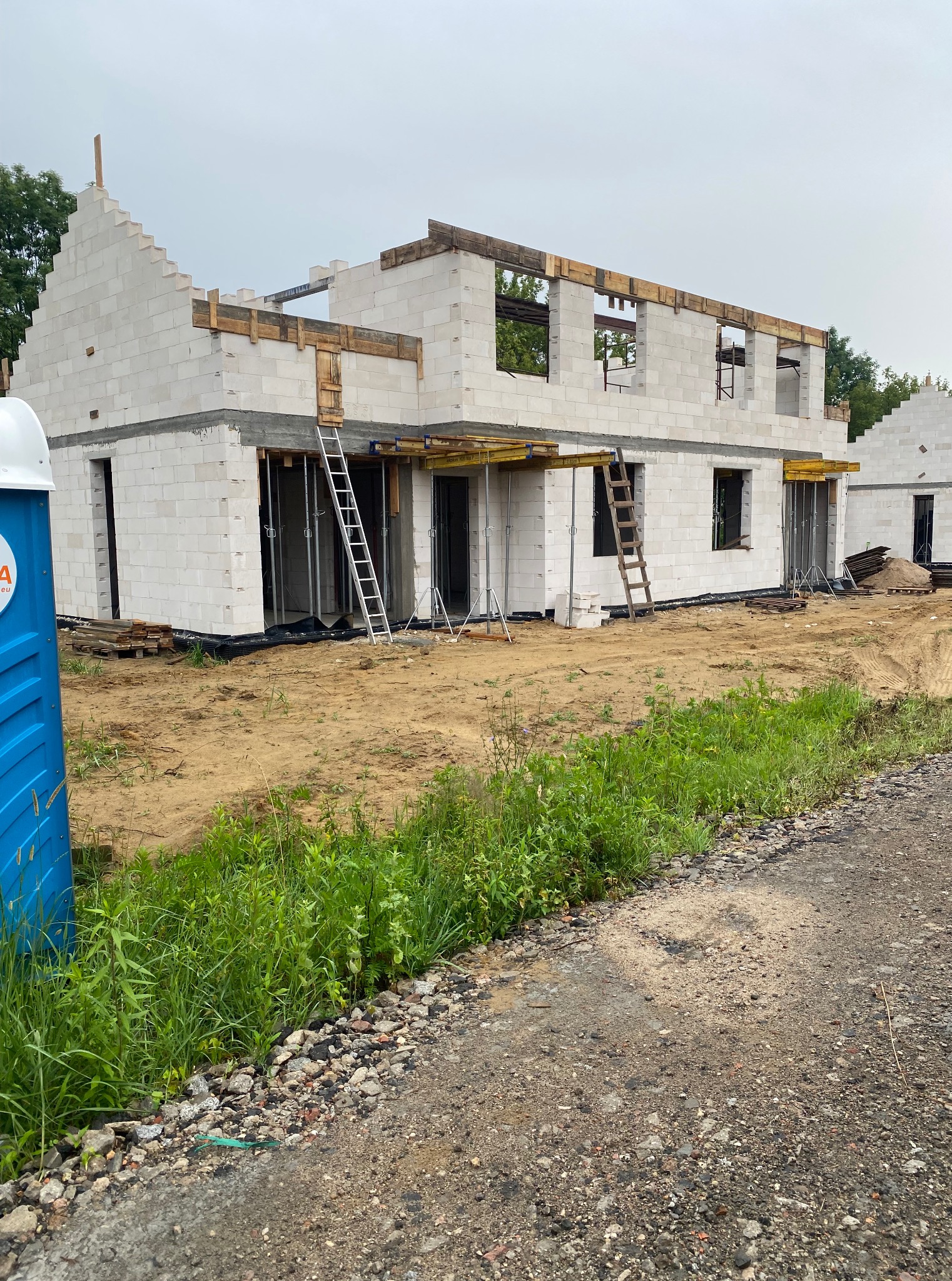 Budowa domu z białych bloczków, widoczne rusztowania i drabiny, teren budowy z zielenią. Na pierwszym planie przenośna toaleta.