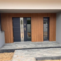 Firma Budowlana EKO-BUD Leszek i Sylwester Wielgosz spółka cywilna - Nowoczesne wejście do domu z szarą stolarką drzwiową i okładziną ścienną imitującą drewno. Ułożona kostka brukowa na podeście. Widoczny młotek.